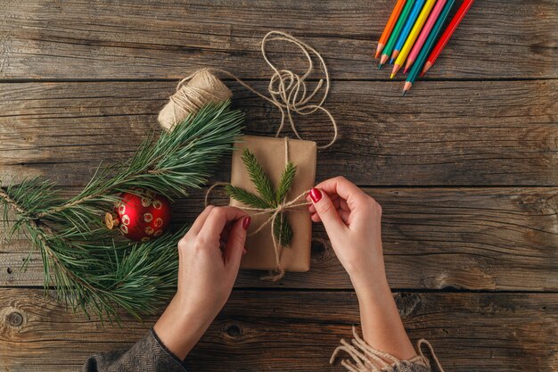 A person wrapping a gift with brown kraft paper and twine, decorated with a sprig of greenery and a small wooden tag. The scene emphasizes the use of natural and recyclable materials for gift wrapping.