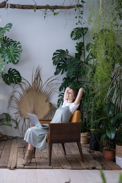 A person sitting in a comfortable armchair, surrounded by a variety of indoor plants in a sunlit room. The scene evokes a sense of calm, relaxation, and connection with nature.