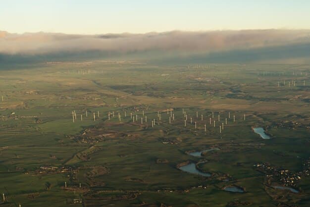 An aerial view of a wind farm in a rural area with rolling hills. Numerous white wind turbines are scattered across the landscape, turning in the wind. The sky is partly cloudy with patches of blue.