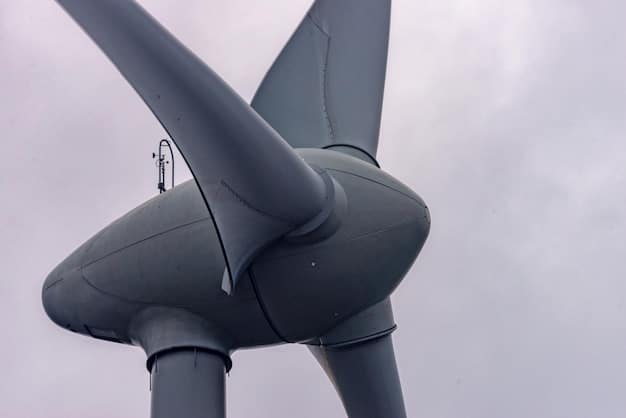 A close-up shot of a wind turbine in operation against a cloudy sky, focusing on the blades turning. The landscape below is green and rolling, emphasizing the integration of renewable energy with nature.