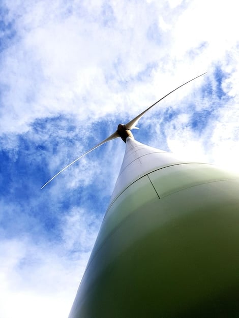 A close-up view of wind turbines turning on a sunny day, with a focus on one turbine in the foreground and several others blurred in the background. The sky is clear and blue.